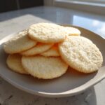 A stack of round, pale yellow sugar cookies coated in sparkling sugar crystals on a neutral plate.
