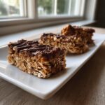 Three homemade protein bars made with oats and topped with drizzled chocolate, resting on a white rectangular plate.