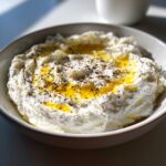 A bowl filled with creamy, textured whipped feta dip, drizzled generously with golden olive oil and cracked black pepper.