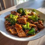 A close-up of saucy chicken and broccoli coated in a rich brown glaze and sprinkled with sesame seeds.