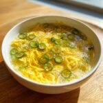 A white bowl filled with bright yellow egg drop soup, topped with sliced green chilies and sesame seeds.