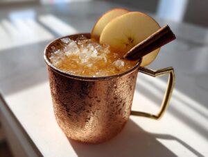 A frosty copper mug filled with an apple cider moscow mule, garnished with apple slices and a cinnamon stick.