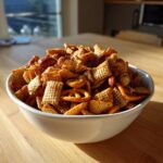 A white bowl overflowing with seasoned chex party mix, including square cereal pieces, pretzels, almonds, and cashews.