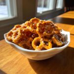 A white bowl filled with glistening, homemade toffee pretzels sprinkled with coarse salt, sitting on a sunny wooden table.