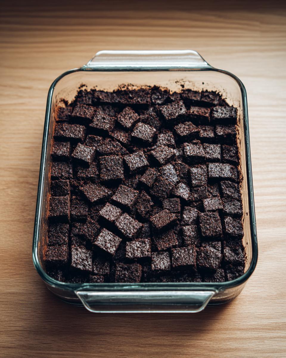 Cubes of dark chocolate brownie layered in a glass baking dish for making brownie refrigerator cake.