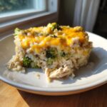 A square serving of cheesy chicken and rice casserole topped with melted cheddar cheese and broccoli florets.