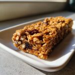 Close-up of a single, chewy homemade granola bar packed with oats, nuts, and dried fruit, sitting on a white dish.