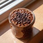 A jar of rich chocolate overnight oats topped generously with mini chocolate chips, sitting in sunlight.