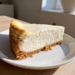 A thick, creamy slice of classic cheesecake recipe on a white plate, showing a graham cracker crust.