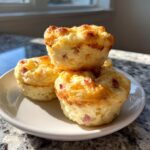 Four golden brown cottage cheese egg cups stacked on a white plate, featuring visible pieces of ham or bacon.