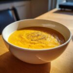 A close-up of a white bowl filled with creamy, vibrant orange cheese soup, topped with freshly ground black pepper.