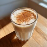A glass of creamy coquito topped generously with ground cinnamon, sitting on a wooden surface.