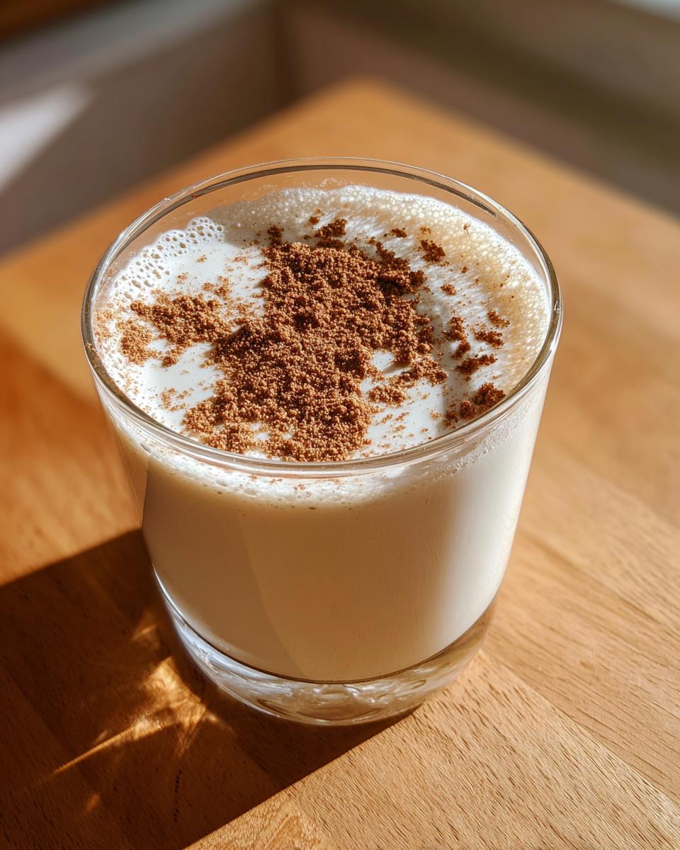 A glass of creamy coquito topped with a generous dusting of ground cinnamon, sitting on a wooden surface.