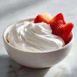 Close-up of a white bowl filled with creamy fruit dip, topped with fresh strawberry slices.