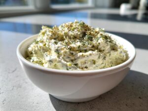 A close-up of creamy pickle dip topped generously with fresh dill and spices, served in a white bowl.
