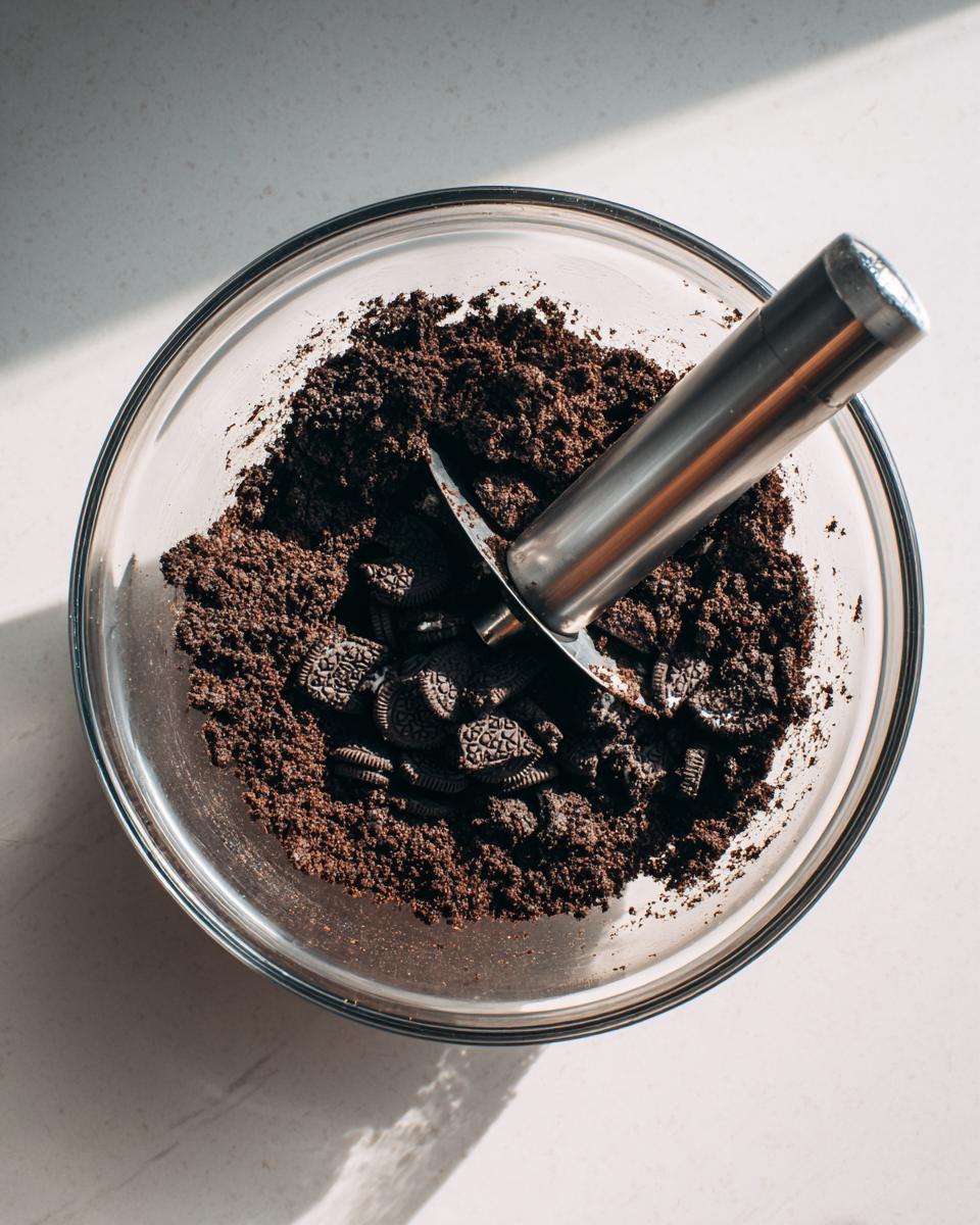 Overhead view of crushed Oreos and whole cookies in a glass bowl being mixed with a metal scoop for oreo dirt cake.