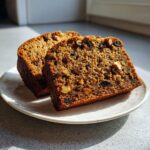 Two thick slices of ultra-moist date nut bread, rich in visible nuts and raisins, resting on a white plate.