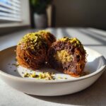 Three decadent dubai chocolate balls, one cut open to show a creamy center, coated in crispy pastry and topped with crushed pistachios.
