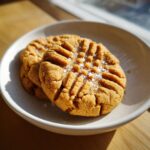 Two classic peanut butter cookies with fork crosshatch marks, topped with flaky sea salt, served on a small white plate.