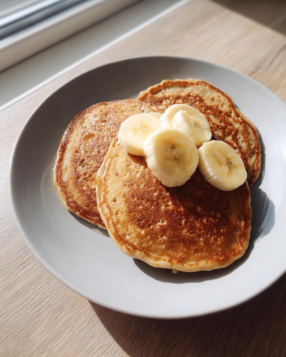 Three fluffy banana oatmeal pancakes stacked slightly on a grey plate, topped with fresh banana slices.
