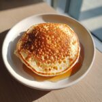 A close-up of fluffy banana pancakes stacked on a plate, drizzled with golden syrup in bright sunlight.