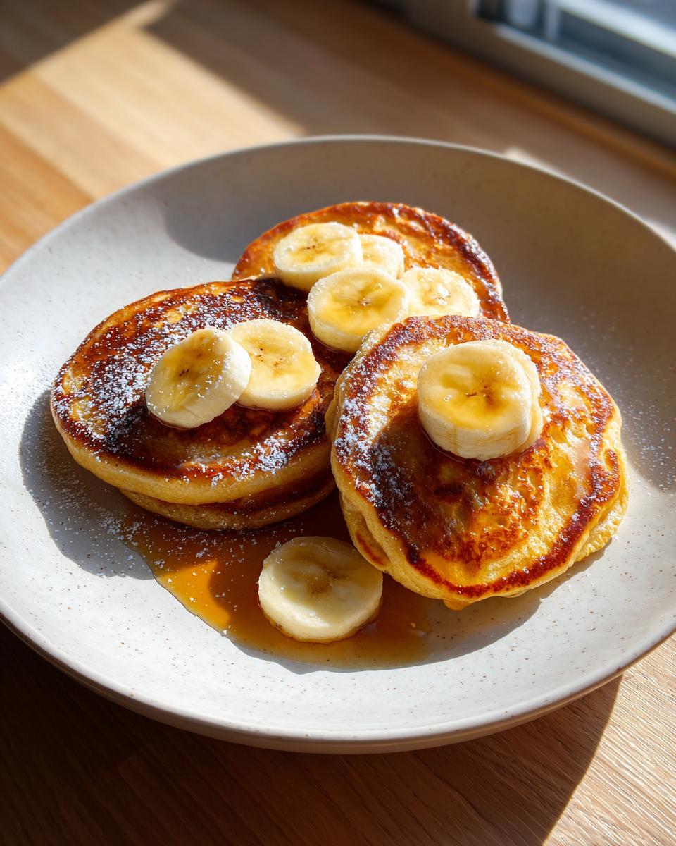 Three golden brown banana pancakes stacked slightly, drizzled with syrup and topped with fresh banana slices and powdered sugar.