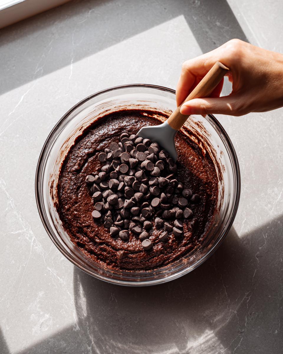 A hand adds chocolate chips to rich, dark batter for sweet potato brownies in a clear glass bowl.
