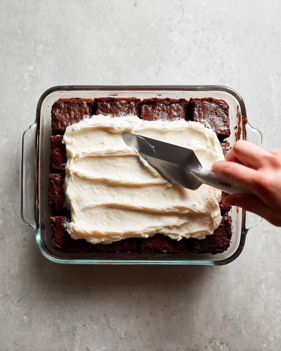 A hand spreads thick white frosting over squares of rich chocolate brownie refrigerator cake in a glass baking dish.
