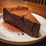 A rich, dark slice of flourless chocolate torte dusted heavily with cocoa powder, served on a light plate.