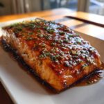 A perfectly cooked, thick fillet of salmon covered in a rich, glossy brown glaze and sprinkled with chopped green herbs.