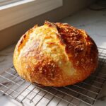 A perfectly baked, round loaf of french bread cooling on a wire rack, featuring a deeply golden, blistered crust.