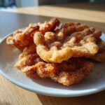 A stack of freshly fried, golden brown buñuelos generously coated in sparkling cinnamon sugar on a white plate.