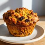 A single, golden-brown morning glory muffin topped with visible raisins and shredded carrots, sitting on a small white plate.