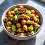 A close-up of a vibrant, fresh bean salad featuring chickpeas, dark beans, chopped cucumber, red pepper, and herbs in a light-colored bowl.