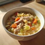 Close-up of a white bowl filled with homemade turkey soup, featuring shredded turkey, pasta, carrots, and celery.