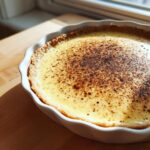 A freshly baked custard pie with a golden, creamy filling dusted heavily with nutmeg, sitting in a white fluted pie dish.