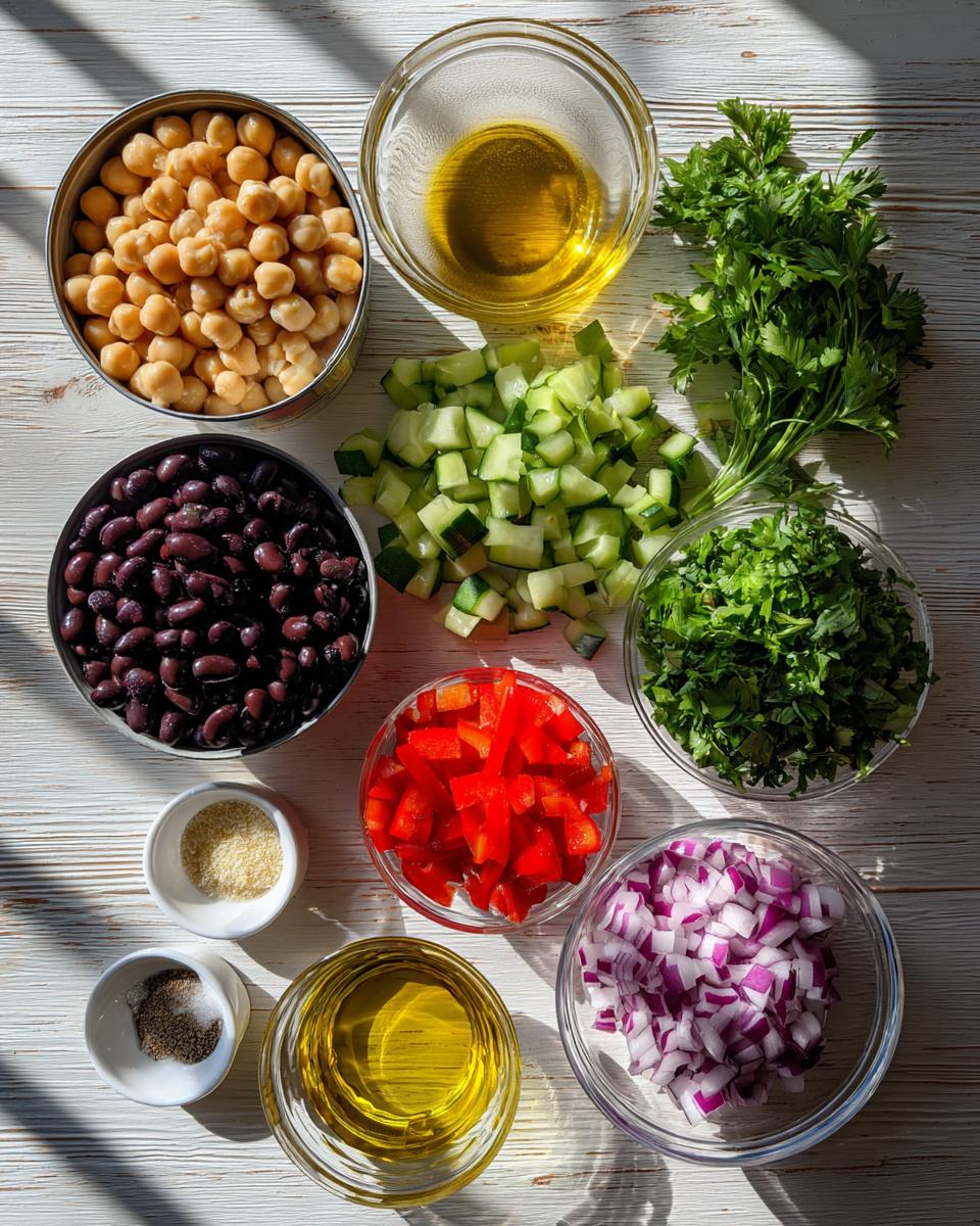 Overhead view of ingredients for bean salad: chickpeas, black beans, chopped cucumber, red onion, red pepper, parsley, oil, and seasonings.