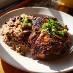 A close-up of a piece of dark, seasoned Jamaican Jerk Chicken served next to rice and peas, garnished with green onions.