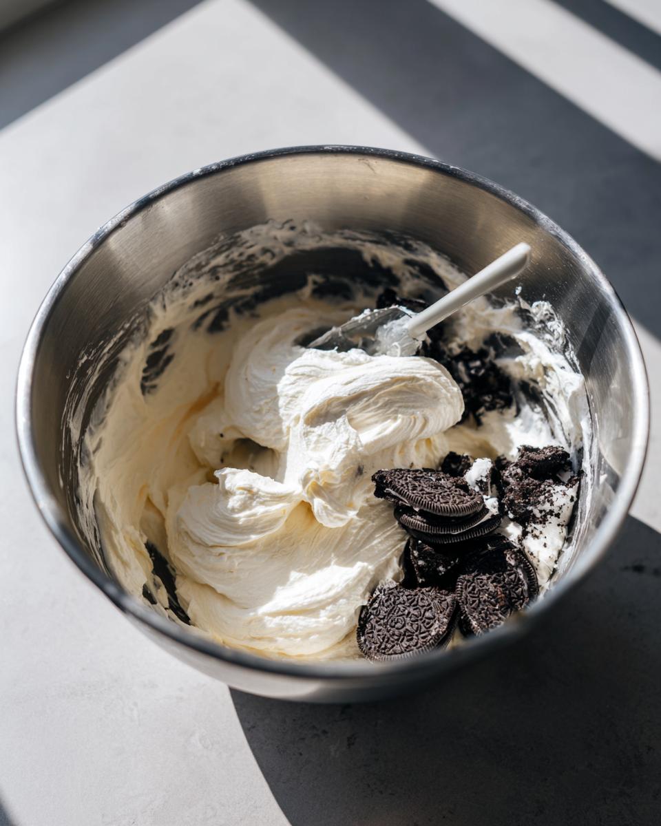 Mixing whipped cream filling with crushed Oreo cookies for an Oreo pie in a stainless steel bowl.