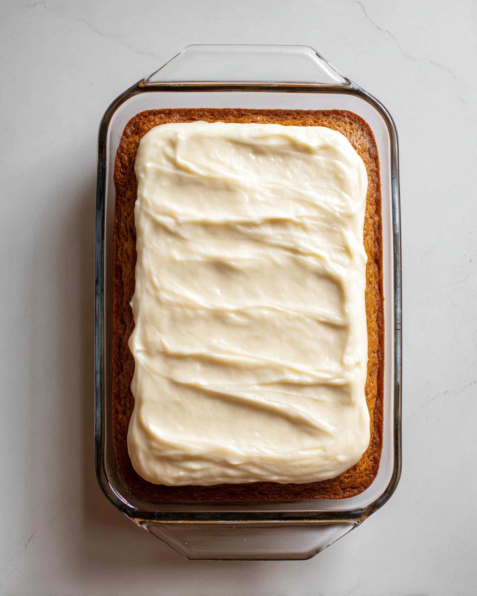 Overhead view of a freshly baked applesauce cake topped with thick, creamy white frosting in a glass baking dish.