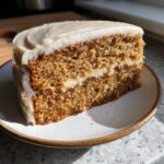 A close-up of a moist slice of applesauce cake layered with light frosting, sitting on a small plate.