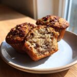 Close-up of two banana nut muffins, one cut in half showing the moist, speckled interior and crunchy nut topping.