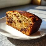 A close-up, sunlit slice of moist greek yogurt banana bread studded with chocolate chips, resting on a white plate.