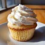 A single, moist vanilla cupcake topped with a tall swirl of white buttercream frosting, lightly dusted with powdered sugar.
