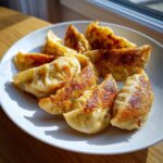 A plate of golden-brown, pan-fried Traditional Chinese Dumplings catching the sunlight.