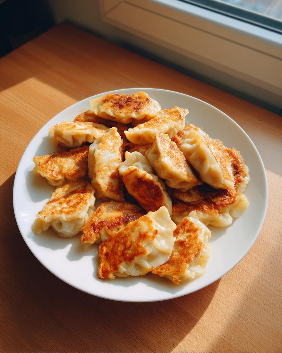 A white plate piled high with golden-brown, pan-fried Traditional Chinese Dumplings sitting on a wooden table near a window.