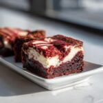 A close-up of a rich, fudgy red velvet cheesecake brownie with a creamy white swirl, served on a white dish.