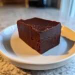 A single, thick square of dark chocolate fudge sitting on a small white plate.
