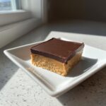 A single square of rich peanut butter bars with a chocolate topping on a white plate.