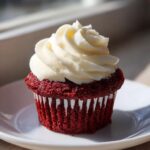 A single, vibrant red velvet cupcake topped with a generous swirl of white cream cheese frosting, sitting on a white plate.